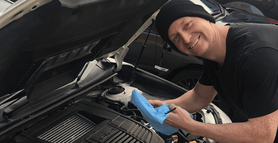 A smiling mechanic wearing a black beanie and t-shirt performs maintenance on a car engine, holding a blue cleaning cloth near the engine compartment.