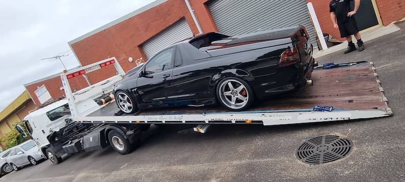 Black Holden Maloo ute being loaded onto a flatbed tow truck outside industrial units, showcasing professional towing service for performance vehicles.