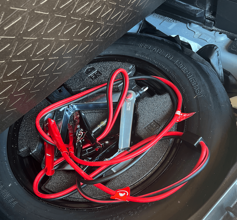 Red jumper cables coiled inside a spare tire well, showing emergency roadside equipment with black diamond-plate surface visible above.