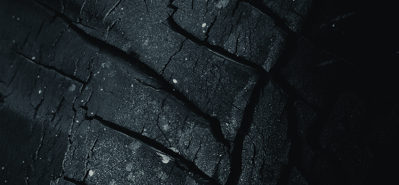 Dramatic close-up of worn car tire treads showing signs of wear and aging, photographed in dark lighting to emphasize texture and grooves.