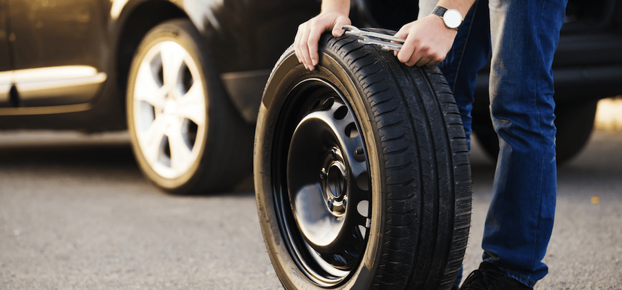 Close-up of a person in blue jeans rolling a spare tire on asphalt, with a parked car visible in the background. The spare tire shows detailed tread pattern and black steel rim.