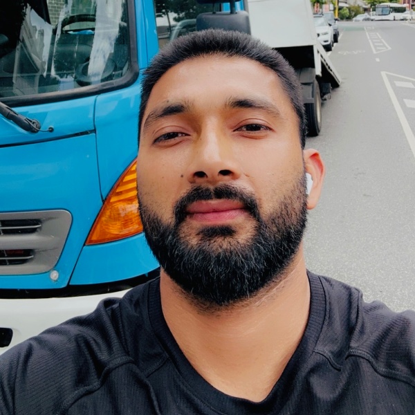 Amritbal Singh, professional tow truck operator at Fast Melbourne Towing, taking a selfie in front of the company's blue tow truck, wearing a black shirt and displaying a friendly, professional demeanour