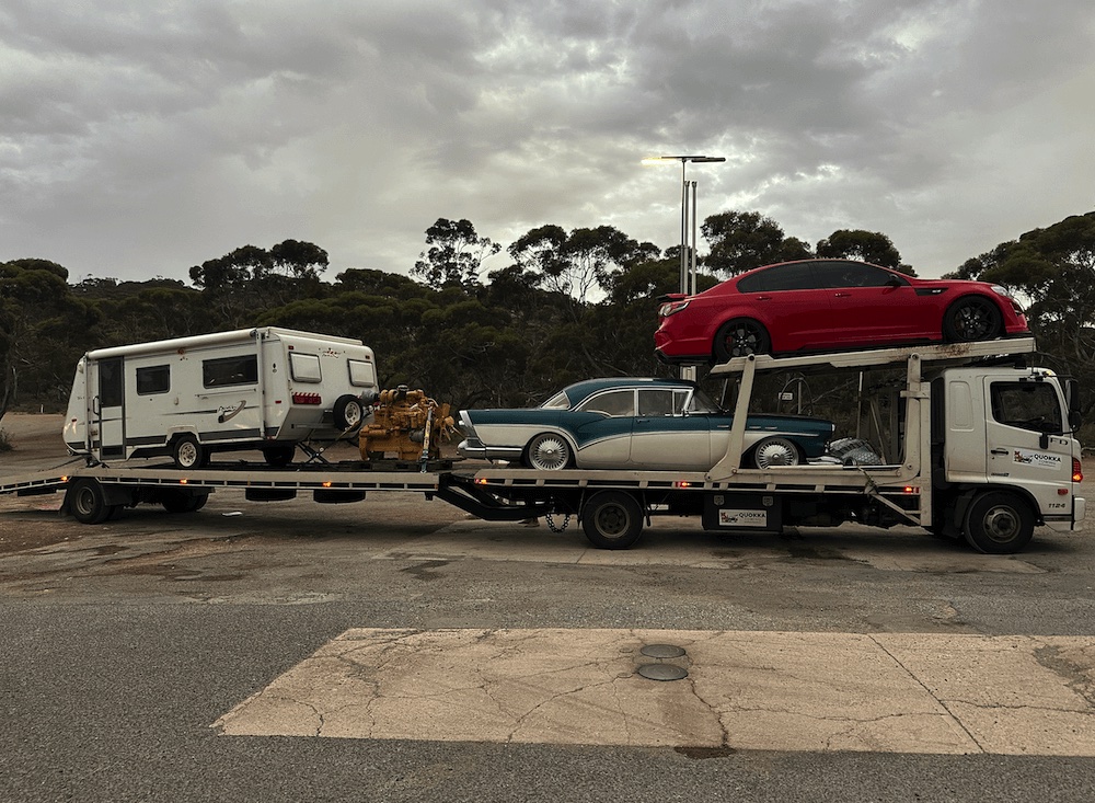 Multi-level tilt tray truck carrying a caravan, a classic car, industrial equipment, and a red sedan