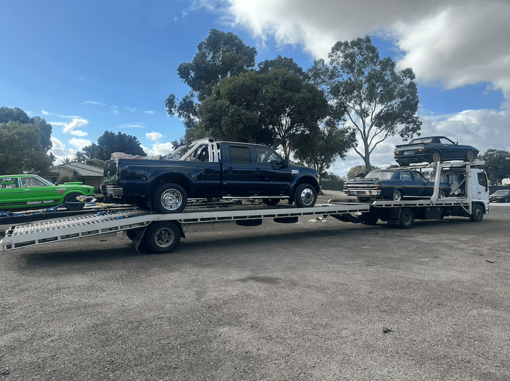 Two car transport trucks loaded with multiple vehicles. The foreground truck carries a navy blue Ford F-series pickup truck, while the second truck in the background is packed with classic cars, including what appears to be vintage sedans. The trucks are parked on a gravel lot with eucalyptus trees visible in the background on a partly cloudy day.